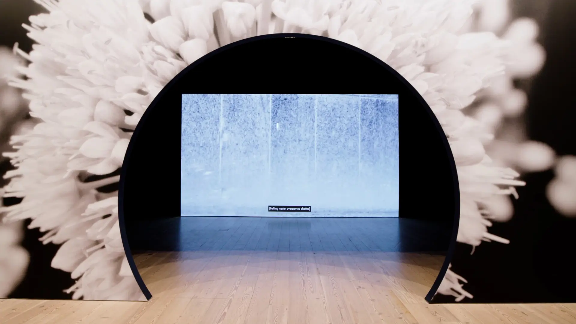 An art installation with a circular archway, flower backdrop, and screen displaying falling water with the caption "[Falli...