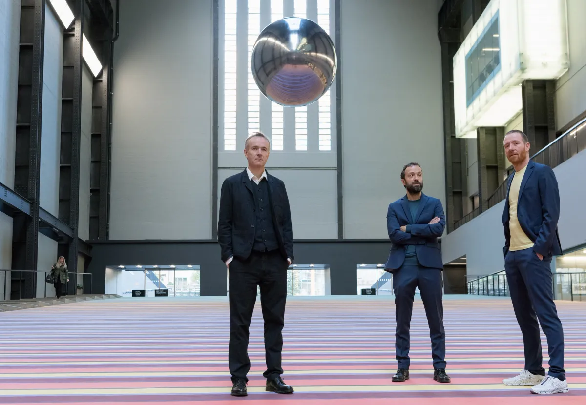 Three individuals standing in a vast gallery space with a reflective sphere hanging above them.