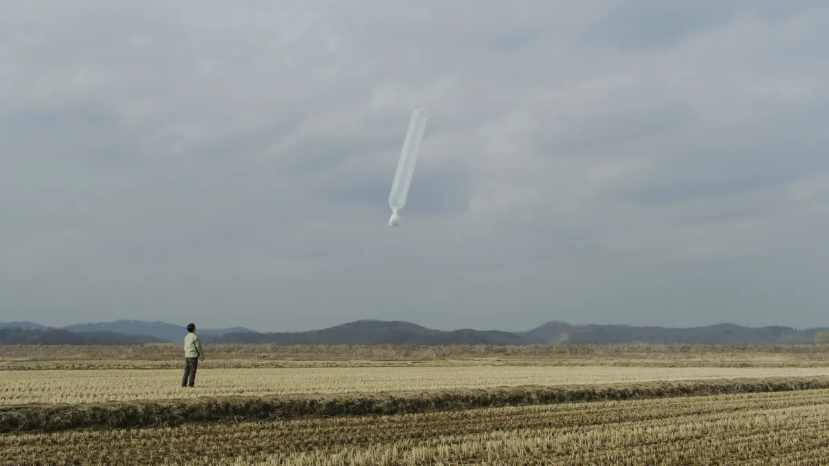 An individual stands in an open field focusing on a floating balloon in the distance.