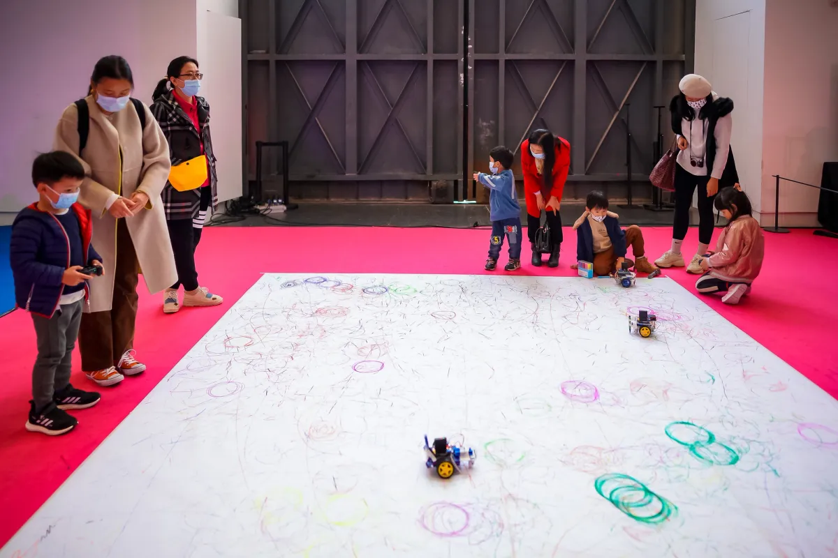A large white surface is surrounded by families. Children operate toy cars on the surface, leaving trails of color, creating various shapes and forms.