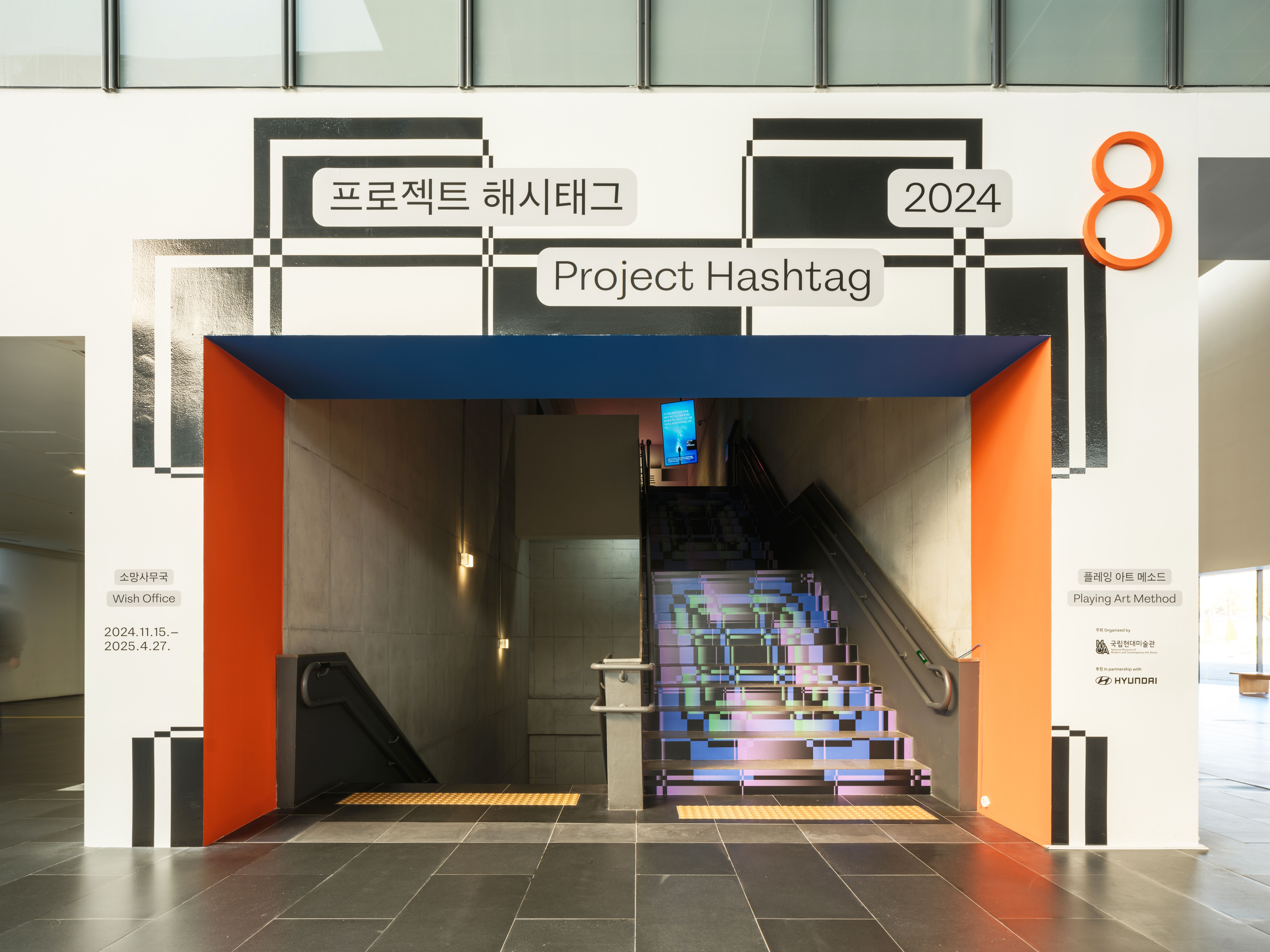 A brightly lit museum entrance featuring a bold architectural frame in orange and blue, leading to a stairwell. The stair ...