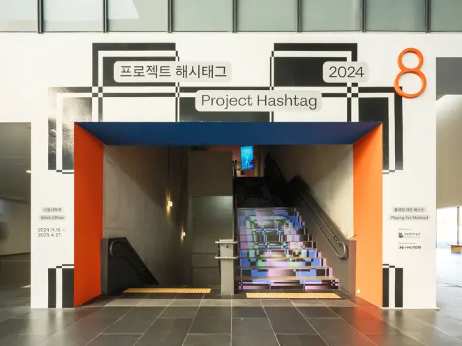 A brightly lit museum entrance featuring a bold architectural frame in orange and blue, leading to a stairwell. The stair ...