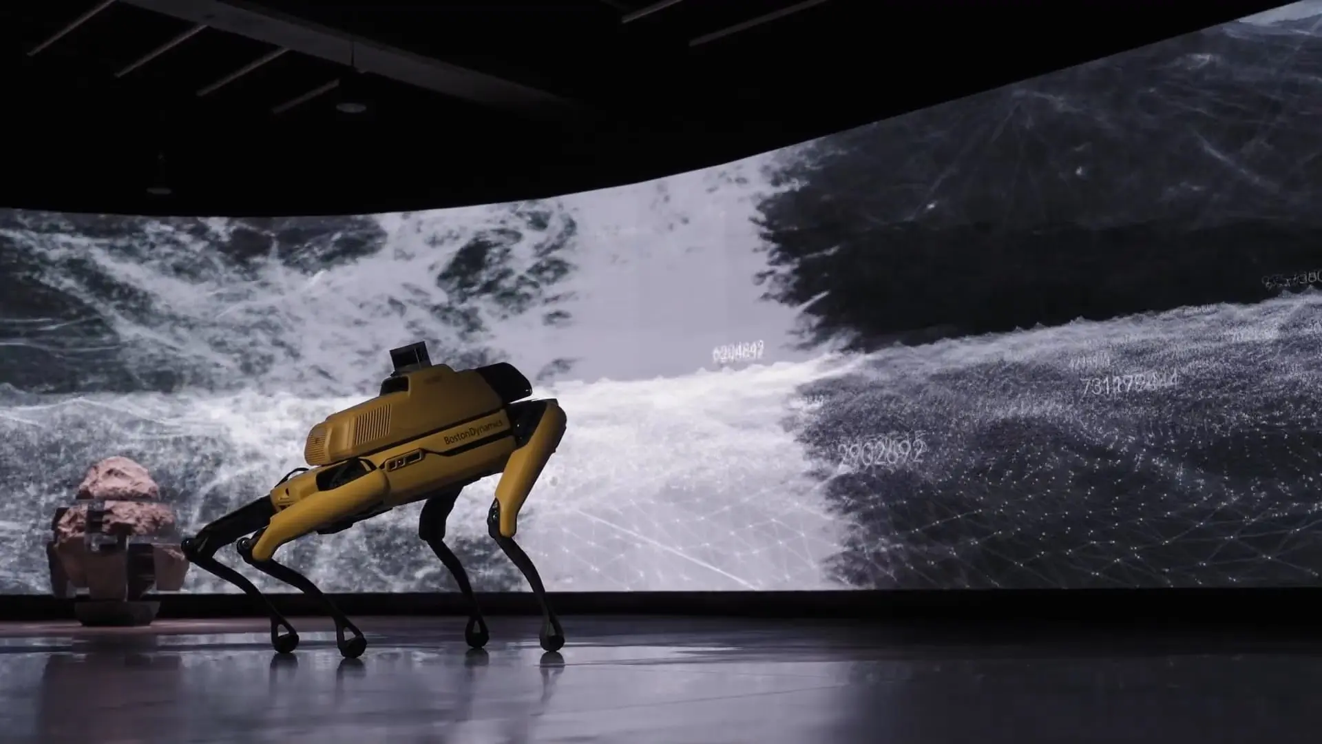 Spot the robot dog moves in front of a large digital screen displaying abstract data and wave patterns.