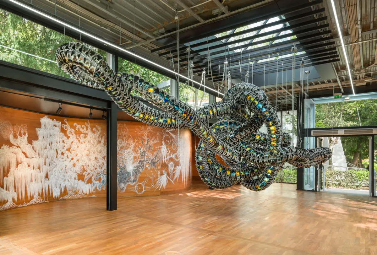 A metallic art installation reminiscent of a snake node hangs from the ceiling in a gallery hall. The hall is surrounded by windows with scenic views of nature.