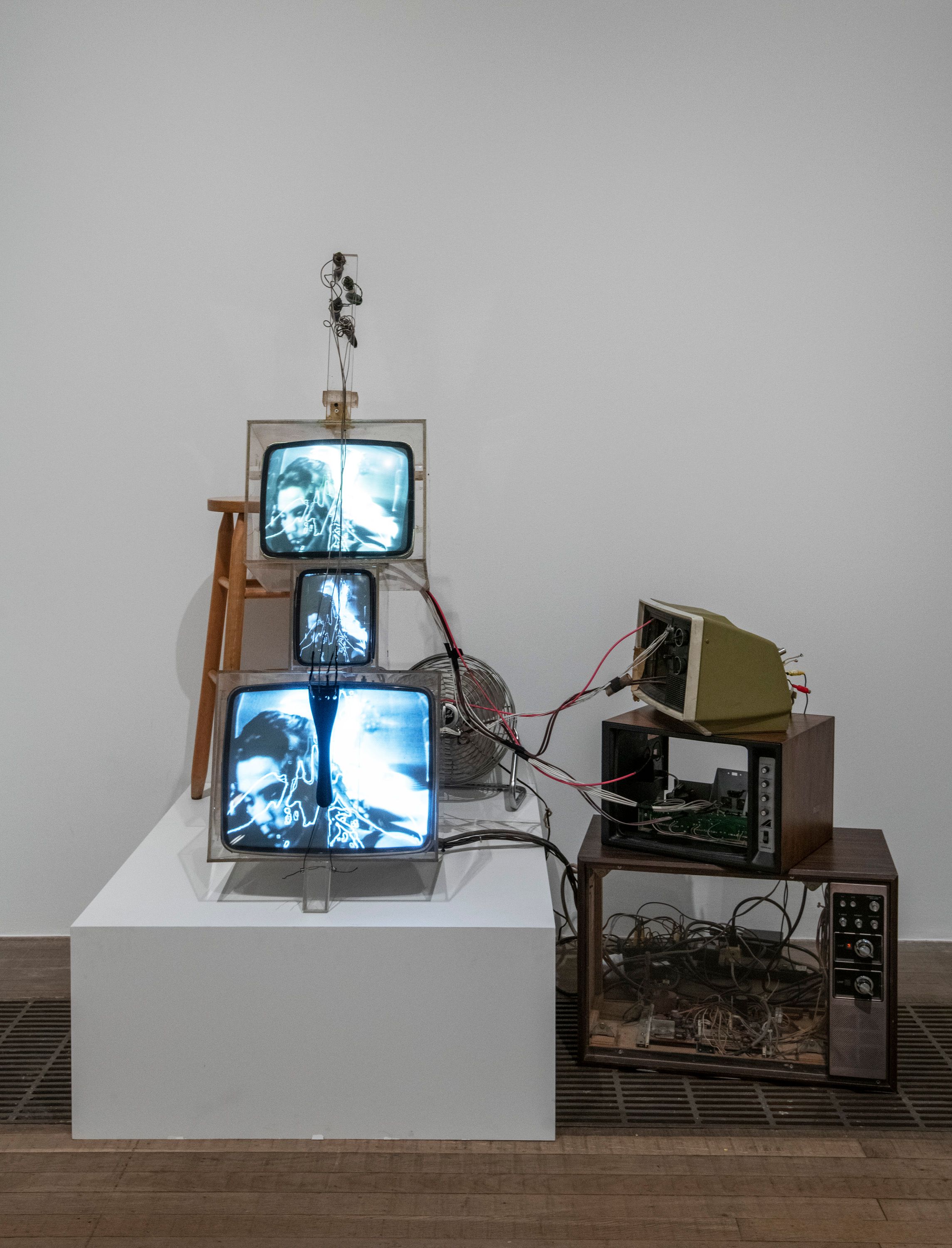 A collection of functioning and broken televisions are arranged haphazardly on a white platform, interconnected by a tangl...