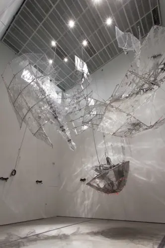 An art installation of transparent rocket-like structures hangs from the ceiling of a well-lit room. A grey rocket nose co...