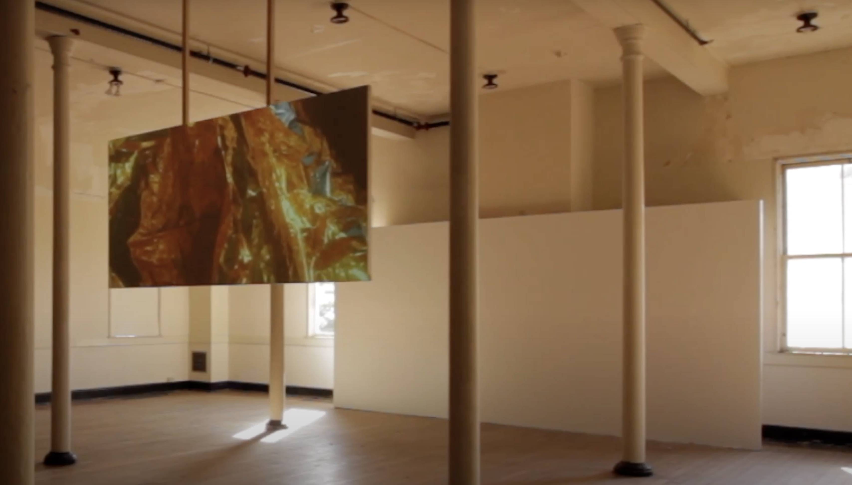 An art piece is suspended from the ceiling in a gallery room adorned with four slim pillars. The room is dimly lit enhanci...