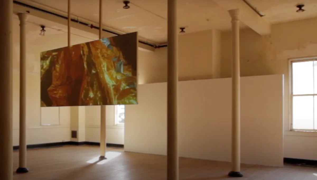 An art piece is suspended from the ceiling in a gallery room adorned with four slim pillars. The room is dimly lit enhancing the projection mapping on the art piece.