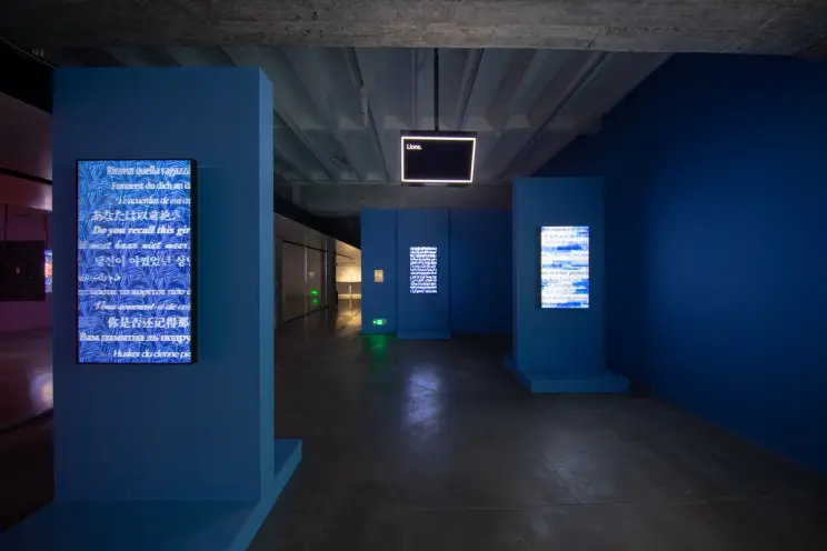 Art exhibit hallway with blue pillars displaying text and overhead neon sign.