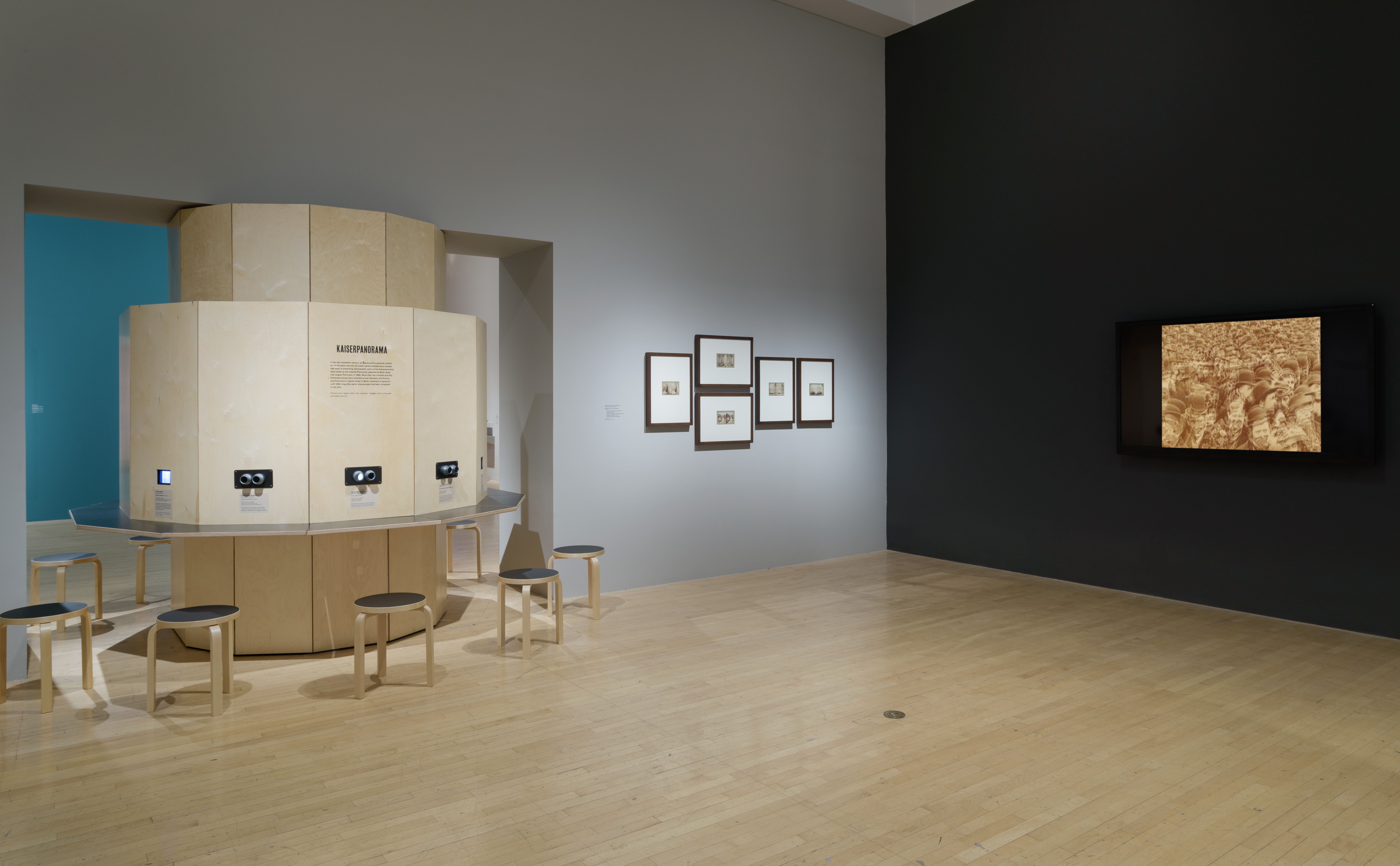A wooden near-circular table with several chairs around it sits in a room with wooden floors. A black wall displays a dese...