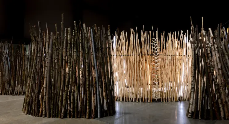 A circular enclosure made of vertically stacked wooden branches glows softly from within in a dark gallery space.