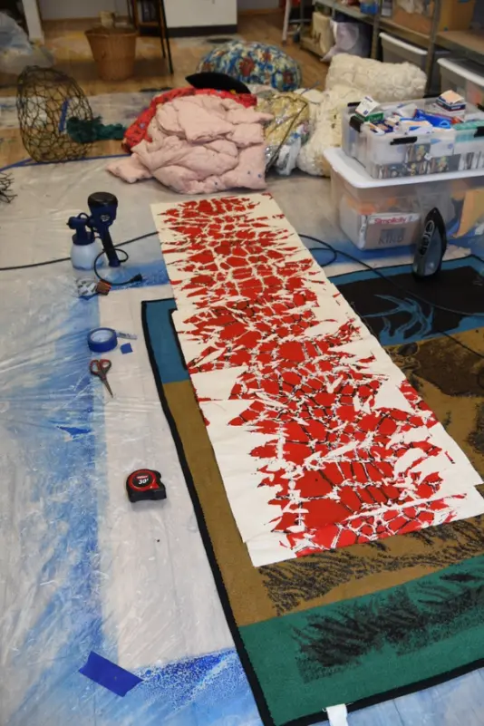 Art studio floor covered in various art supplies and textiles, featuring a distinctive red and white patterned fabric artwork.