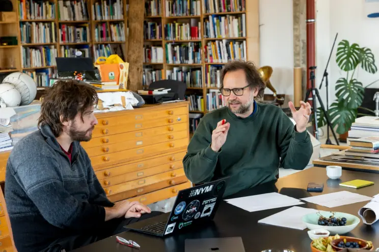 Two individuals engaged in a discussion at a desk cluttered with books and papers in a cozy, book-lined room.