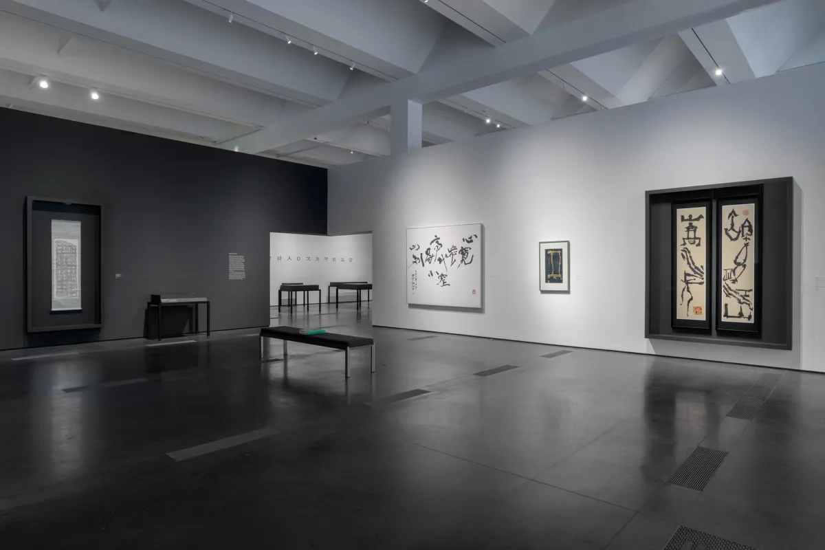 A gallery room with dark flooring displays several art pieces on the walls. A black bench holding a book rests in the center.