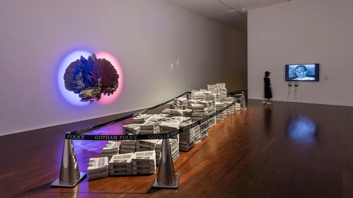 An art installation in a room with wooden floors and white walls, featuring piles of newspapers secured by silver cones and tape labeled "GOTHAM POLICE". In the room, one wall features a screen displaying a blue-tinted face, observed by a standing individual. The opposite wall exhibits a symmetric artwork resembling a brain hemisphere, set against contrasting red and blue backgrounds.