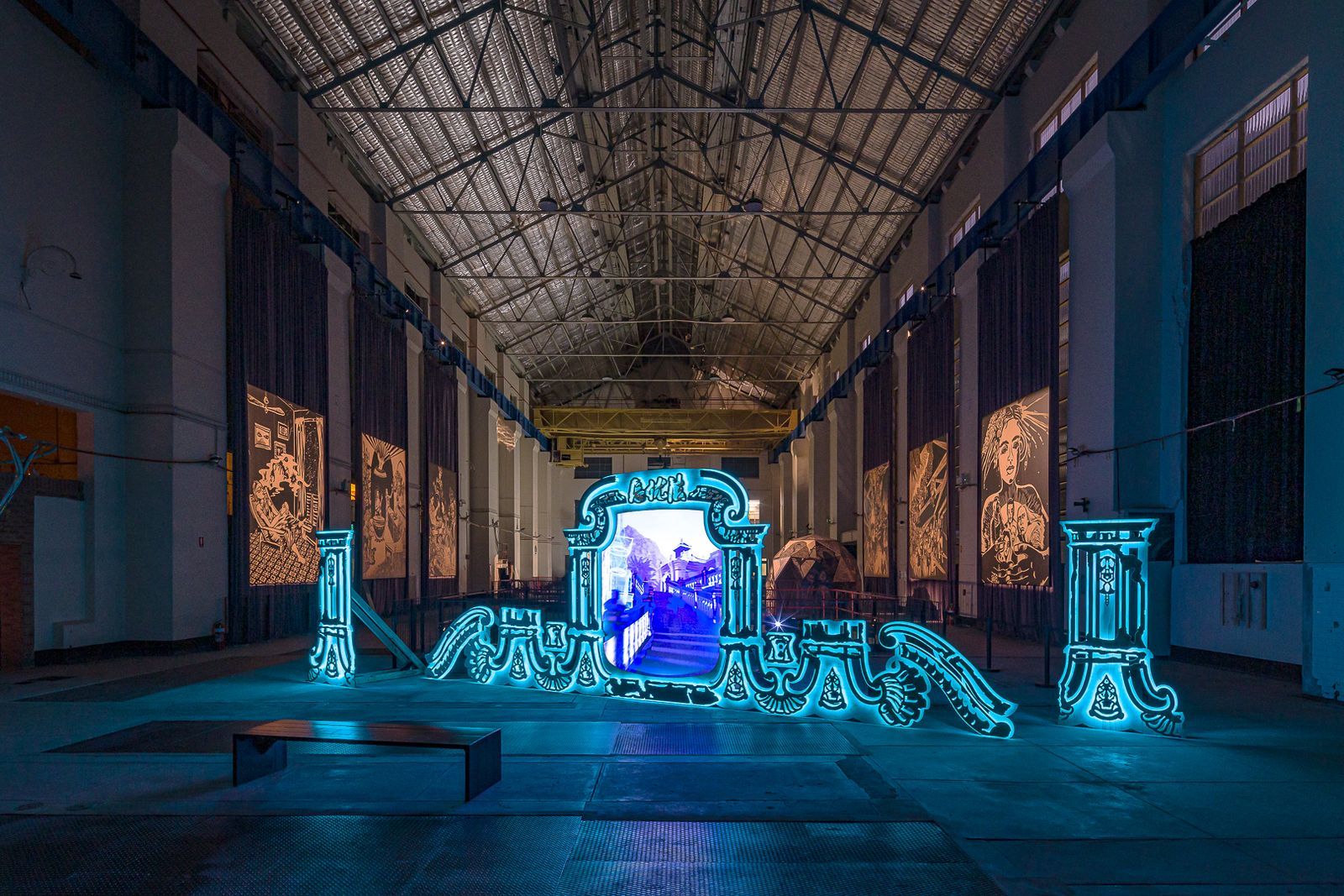 A neon-blue baroque-style frame displaying a video in a vast industrial exhibition hall with large woodcut-style wall hangings.
