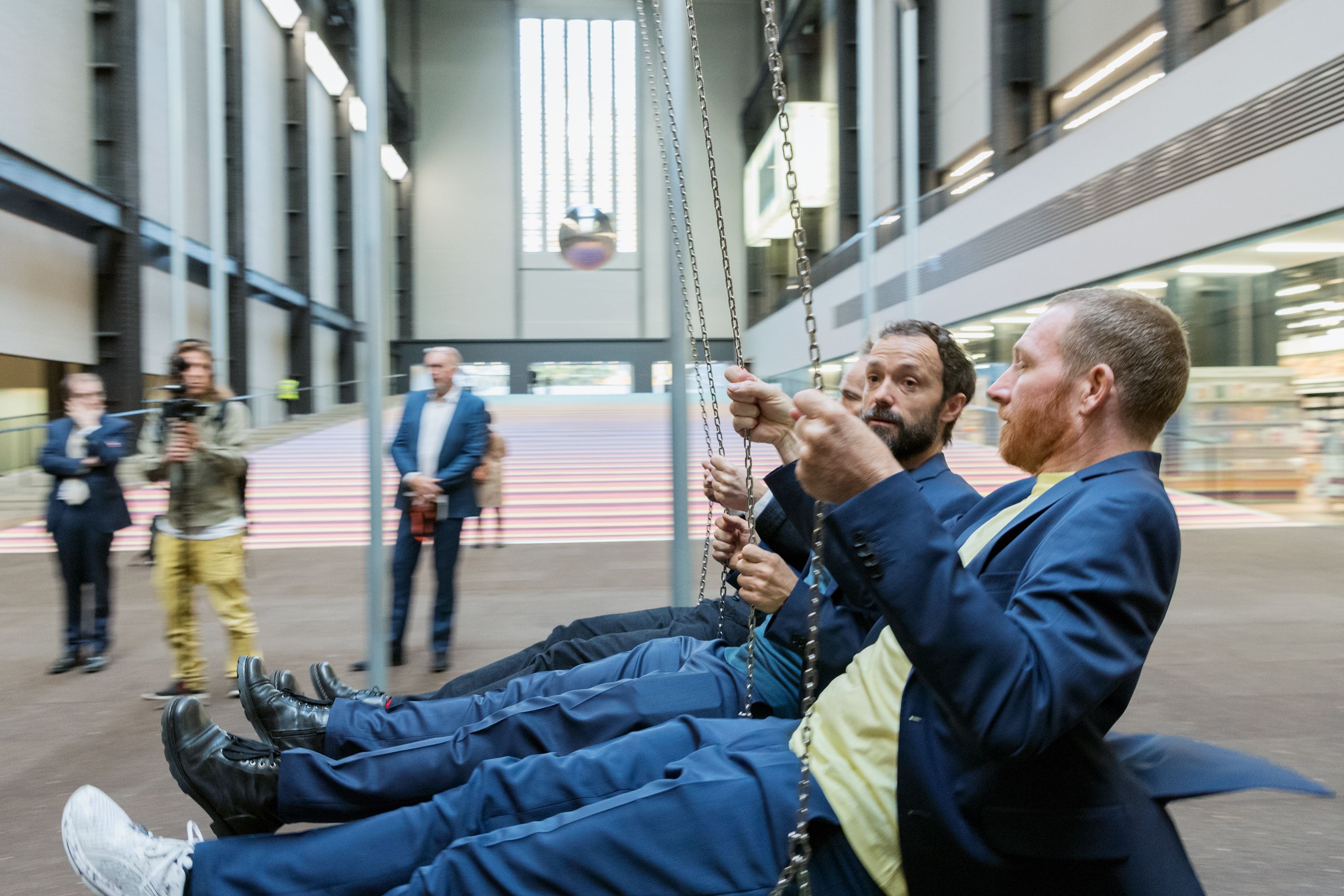 Individuals taking turns on a swinging installation in a spacious art gallery, captured in the midst of their motion.