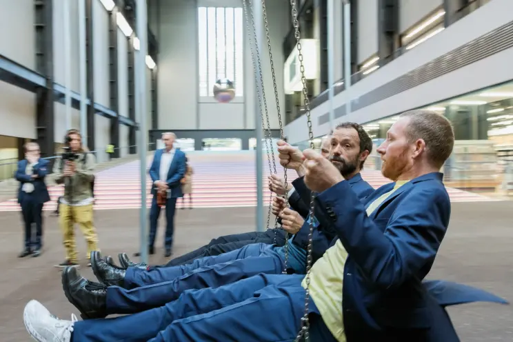 Individuals taking turns on a swinging installation in a spacious art gallery, captured in the midst of their motion.