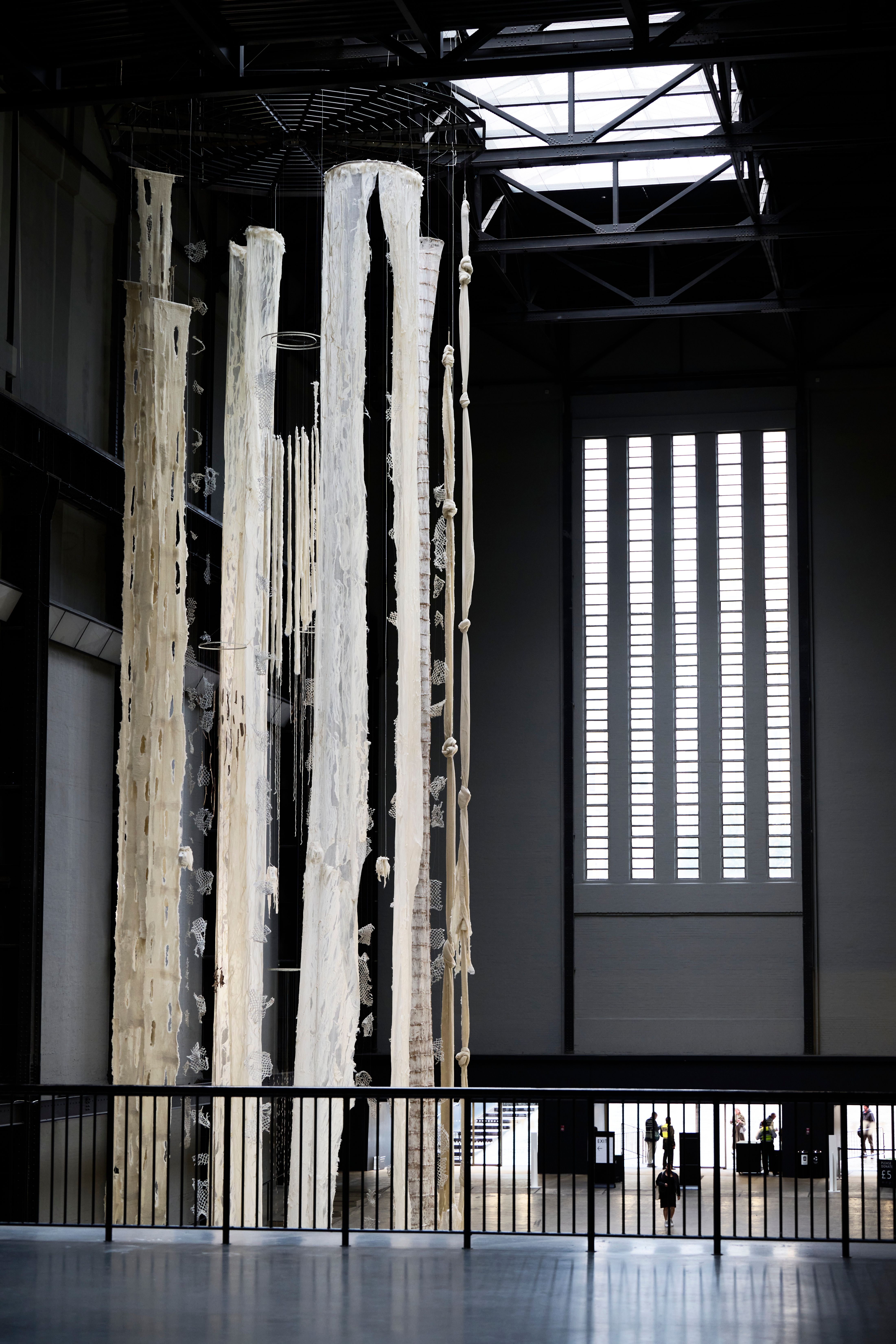 An art installation featuring white torn curtains, fishnets, and rugs suspended from the high ceiling of a gallery hall, v...