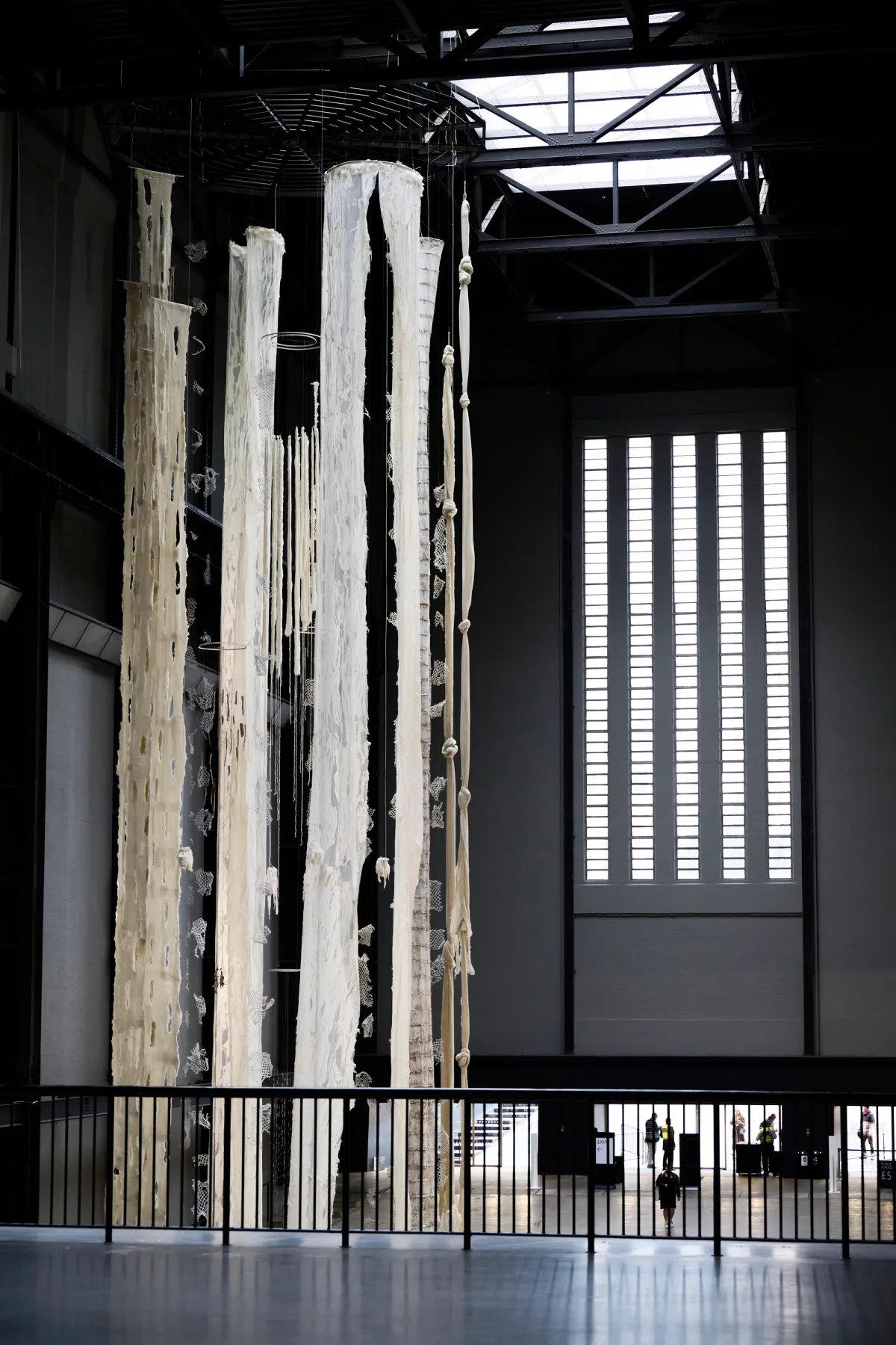 An art installation featuring white torn curtains, fishnets, and rugs suspended from the high ceiling of a gallery hall, viewed from an indoor balcony.
