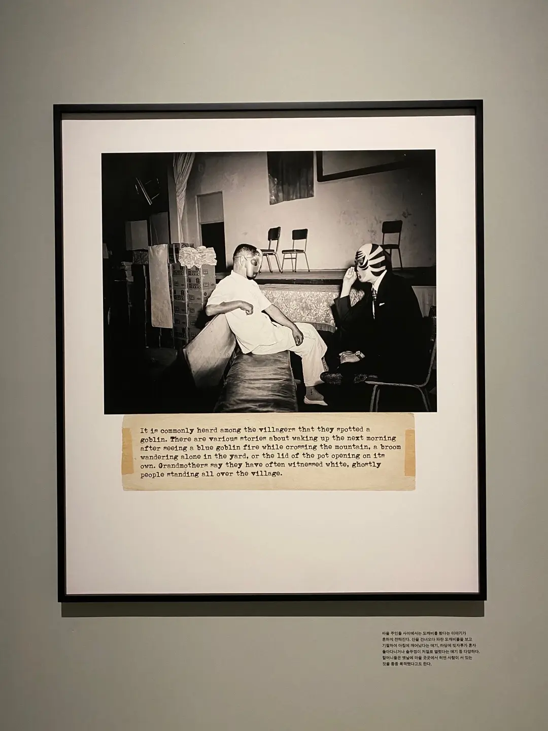 A framed vintage photograph featuring two masked individuals hangs on a gallery wall, accompanied by a textual caption.