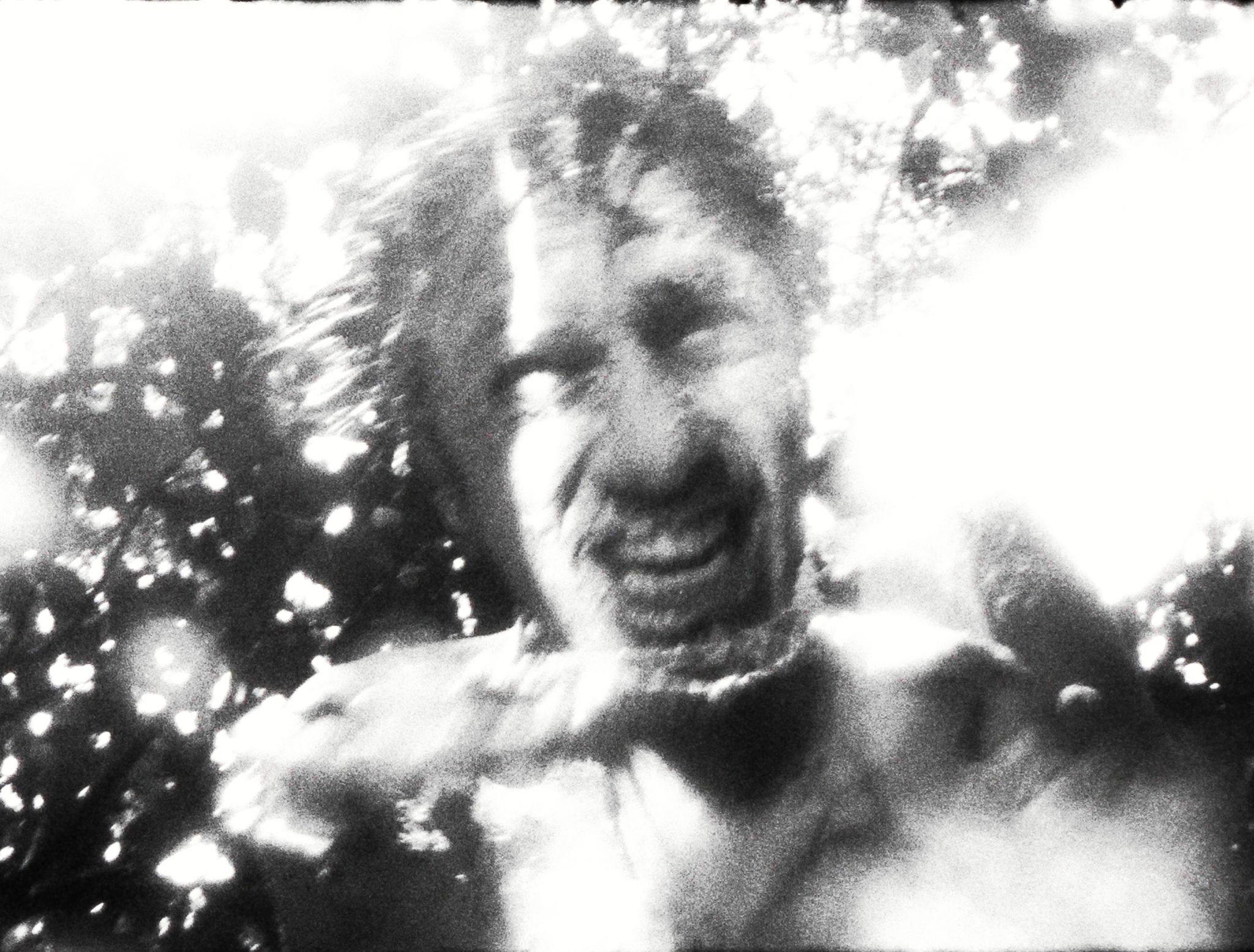 A blurred black-and-white close-up of a figure’s expressive face amid dappled forest light.