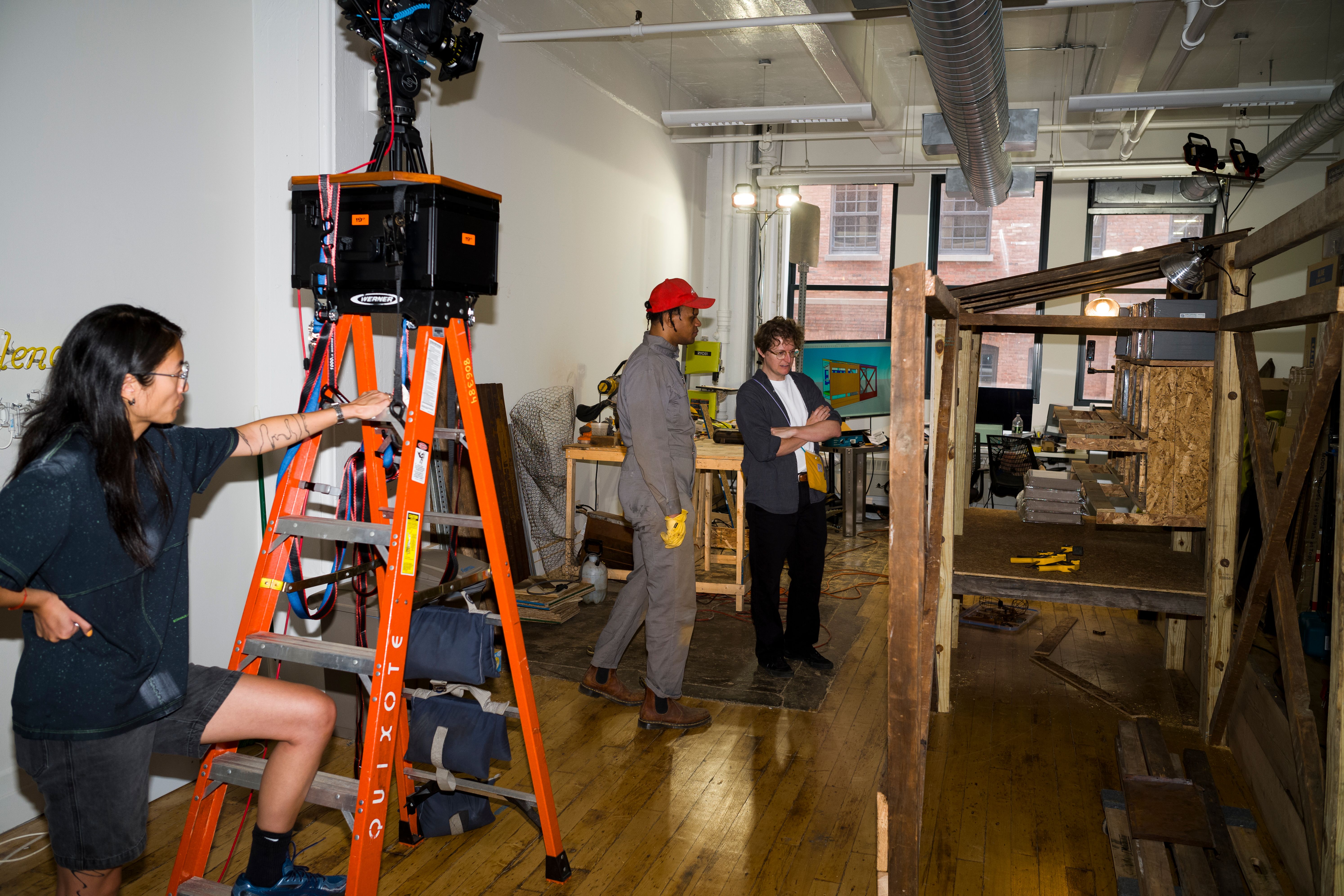 Studio scene with woman on ladder adjusting equipment and two others discussing near woodworking setup.