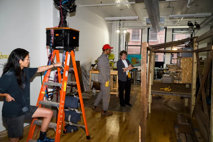 Studio scene with woman on ladder adjusting equipment and two others discussing near woodworking setup.