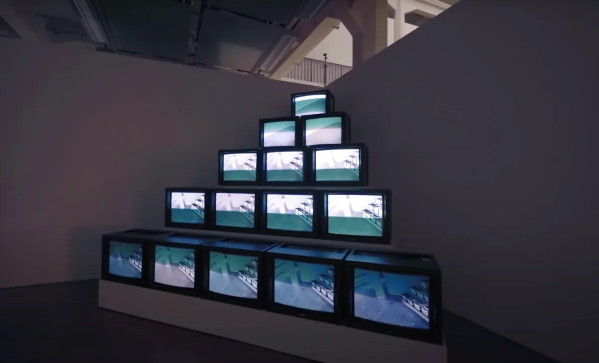 A pyramid-shaped art installation made up of old tv monitors, arranged in descending order of quantity from the base to the peak displayed in a gallery room.