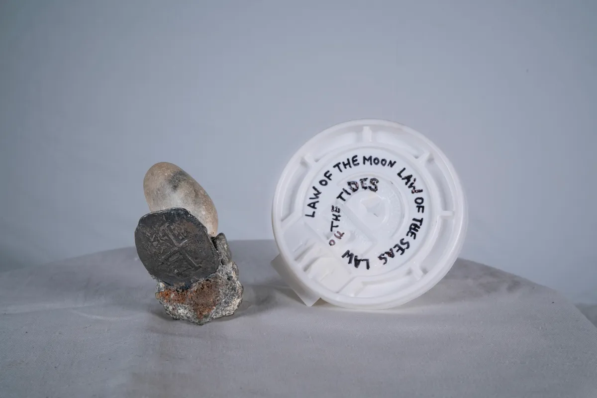 A white round plastic coffee lid with text written on it that reads "Law of the Moon Law of the Seas Law of the Tides" placed on a white background, surrounded by rocks.