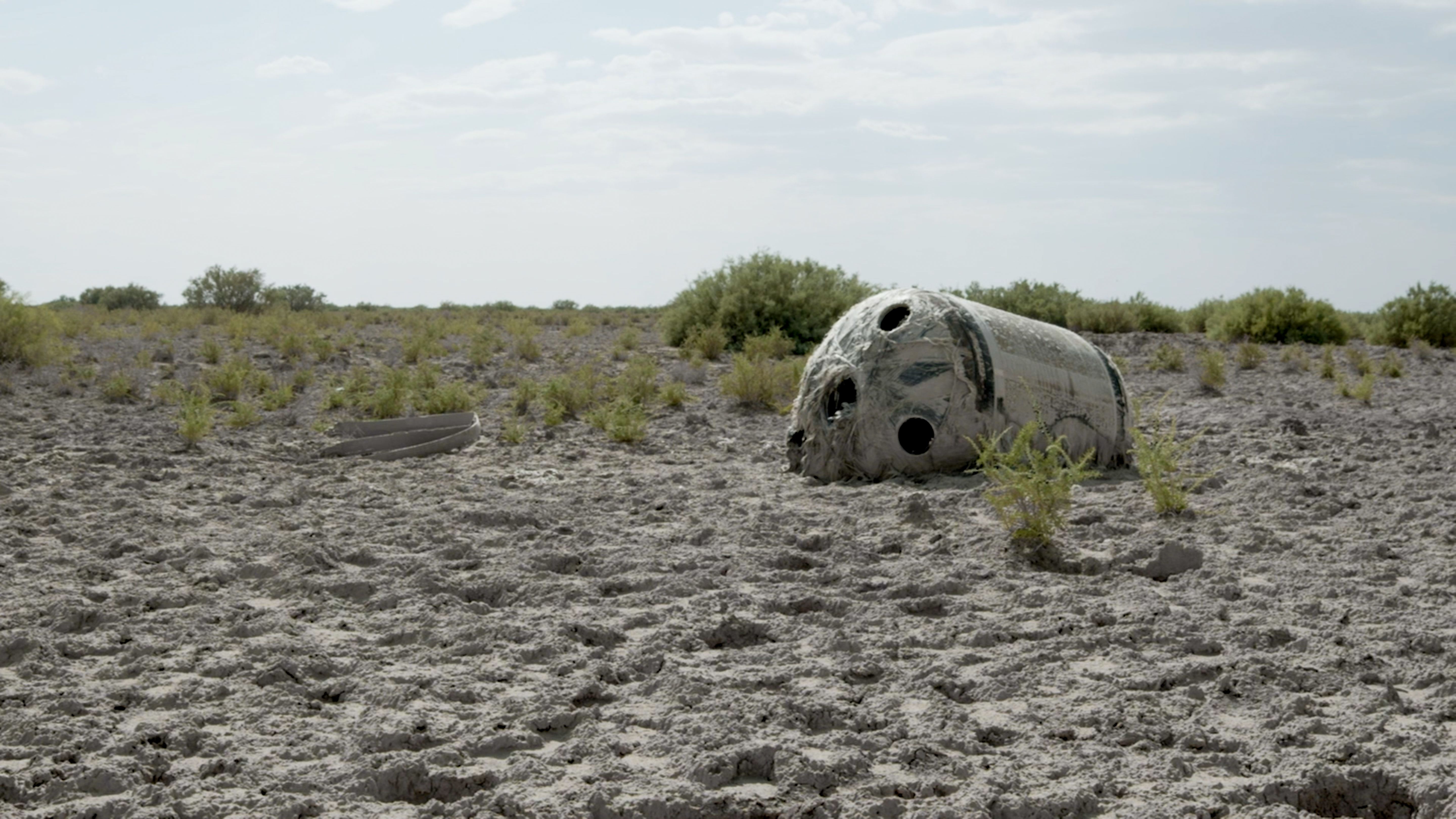 Weathered, capsule-like object lying in a dry, cracked landscape with sparse vegetation.