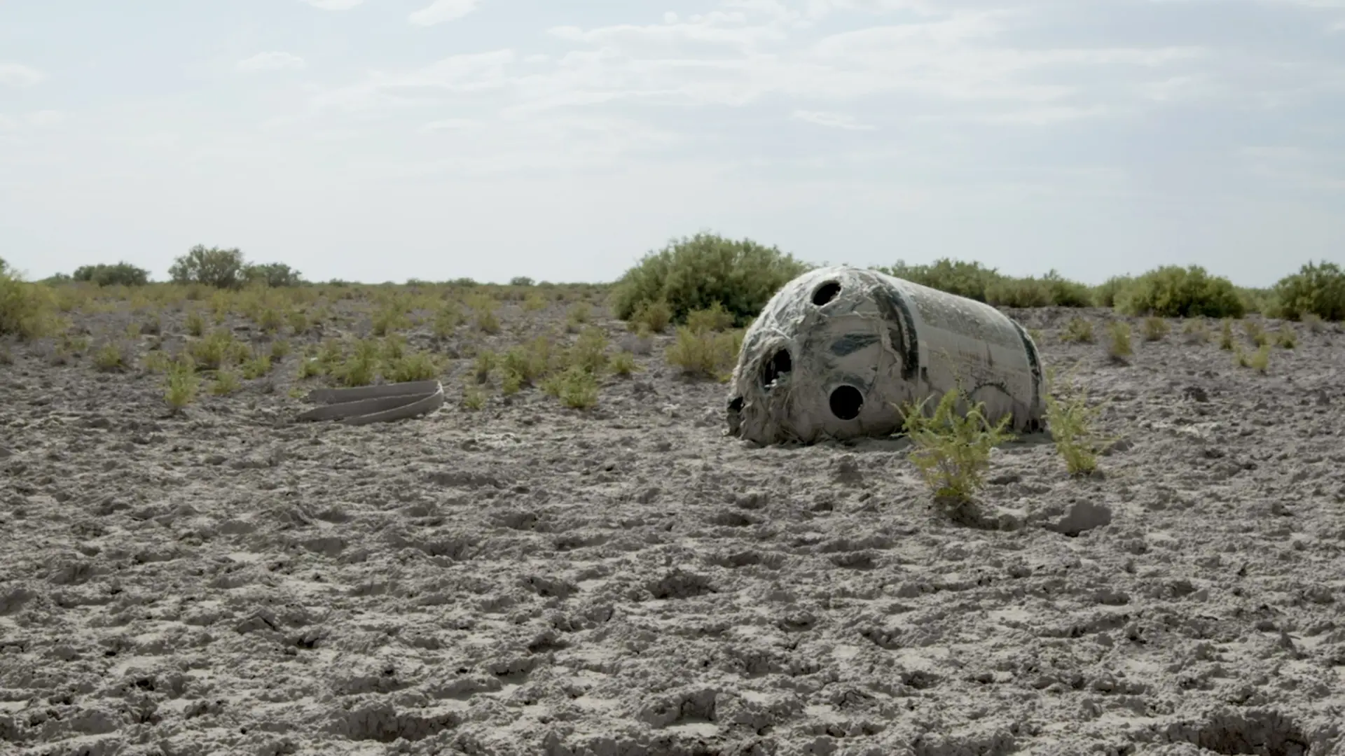 Weathered, capsule-like object lying in a dry, cracked landscape with sparse vegetation.