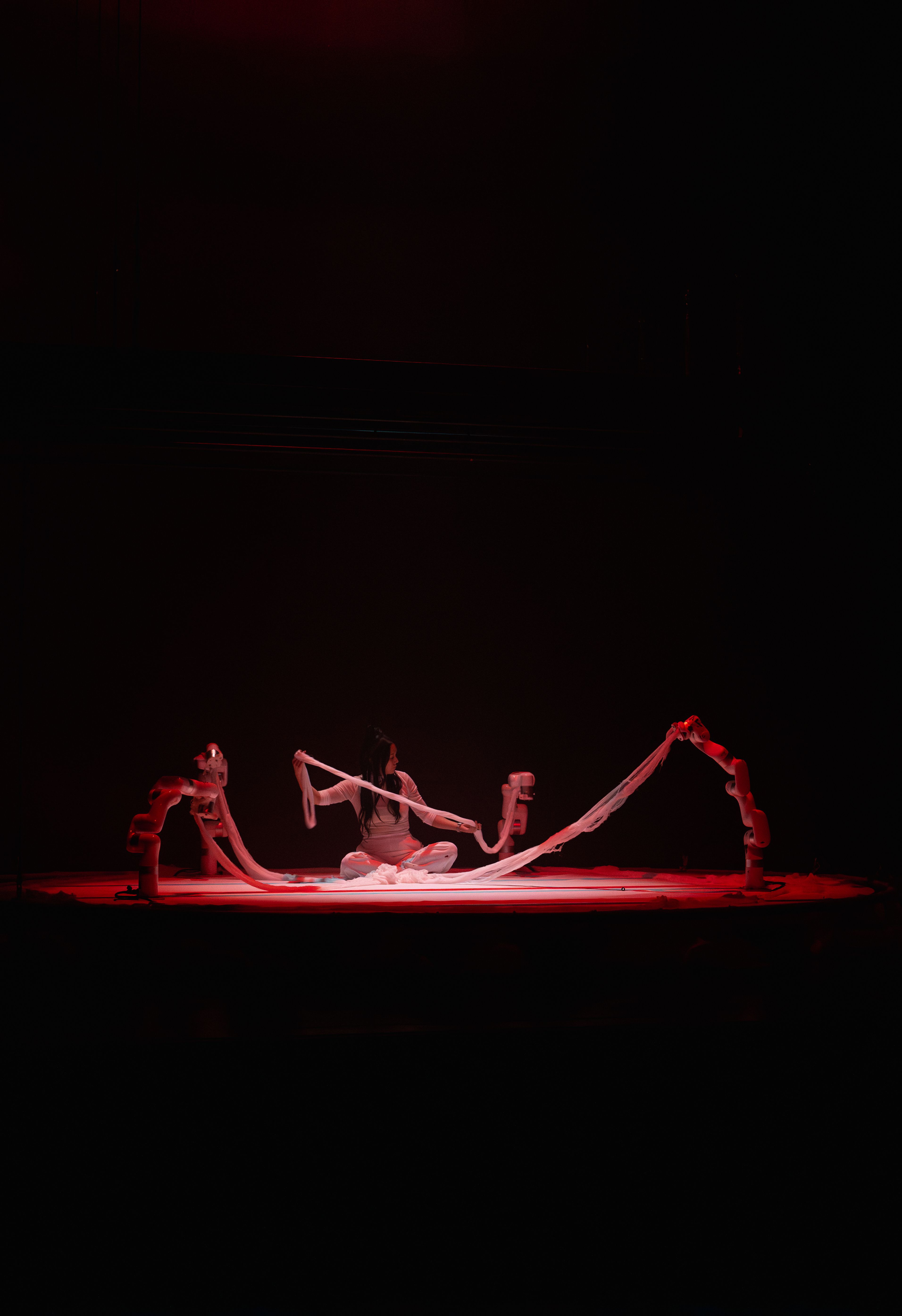 A person dressed in white is seated in the center of a stage under a red spotlight.