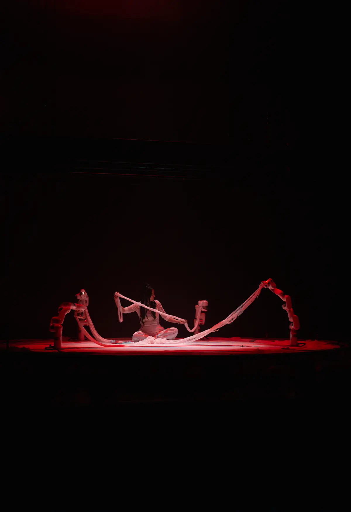 A person dressed in white is seated in the center of a stage under a red spotlight.