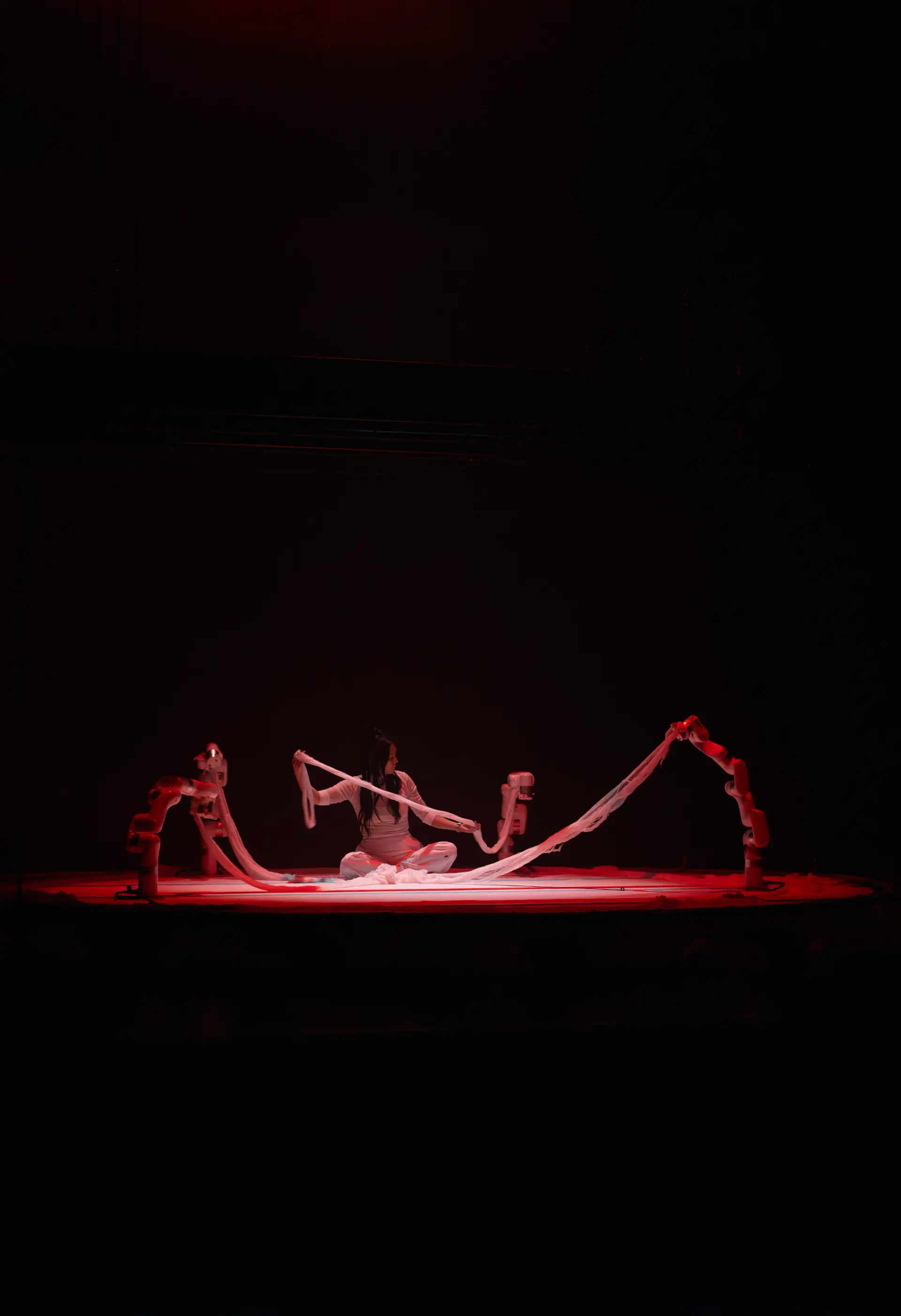 A person dressed in white is seated in the center of a stage under a red spotlight.