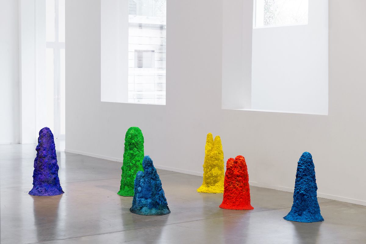 An array of multicolored, textured 3D forms of varying shapes and sizes are scattered on the floor of a pristine gallery s...