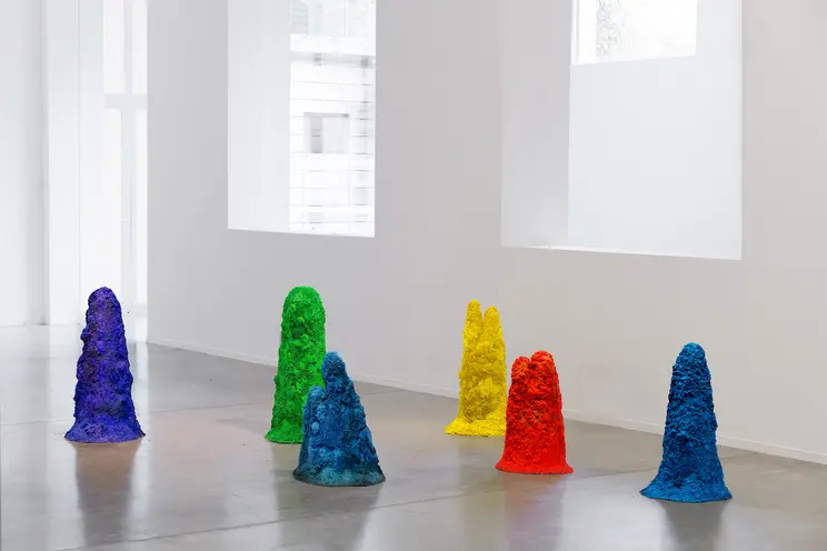 An array of multicolored, textured 3D forms of varying shapes and sizes are scattered on the floor of a pristine gallery s...