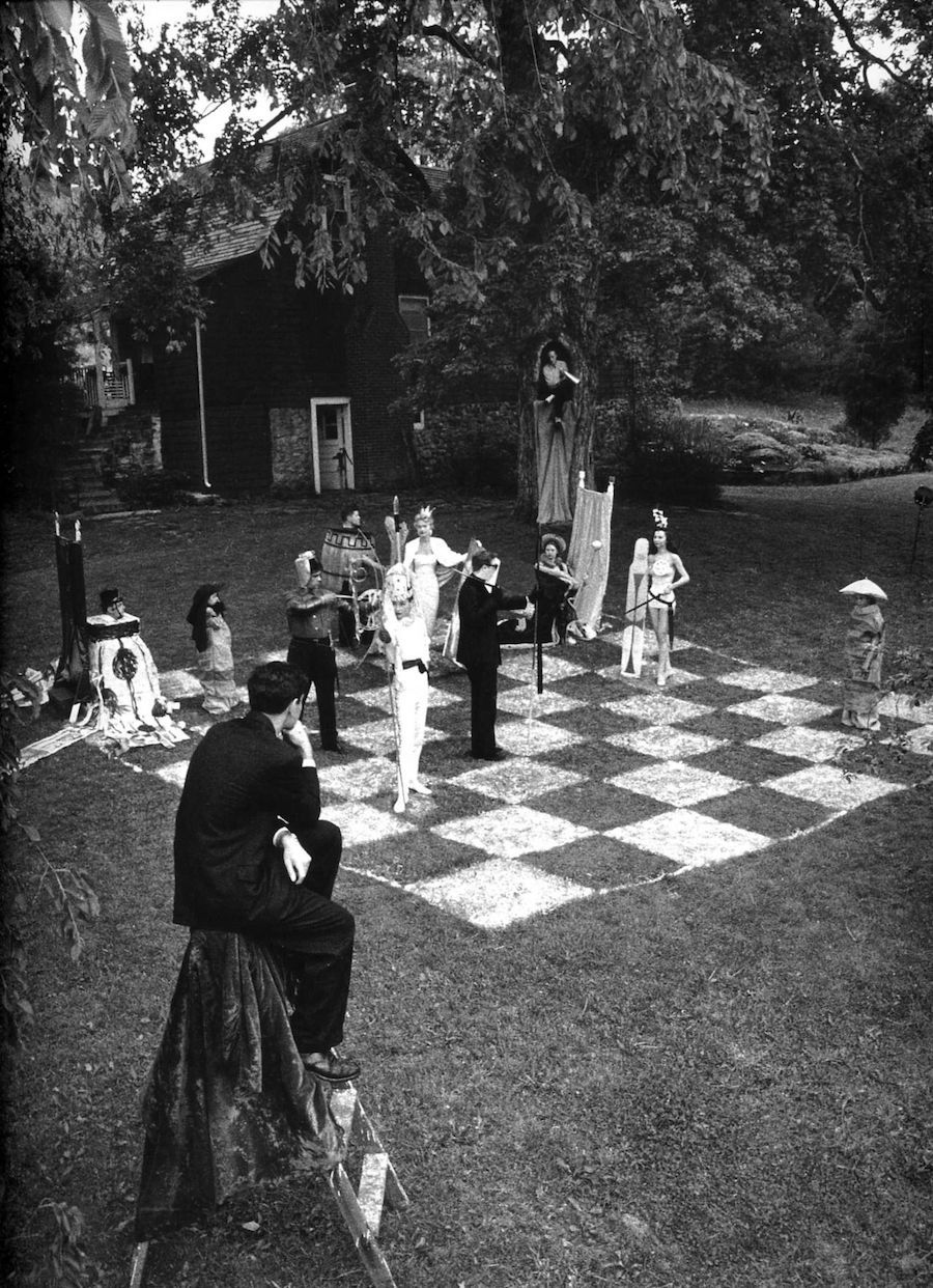 Black-and-white photograph of an outdoor human chess game featuring elaborately dressed participants in a garden setting.