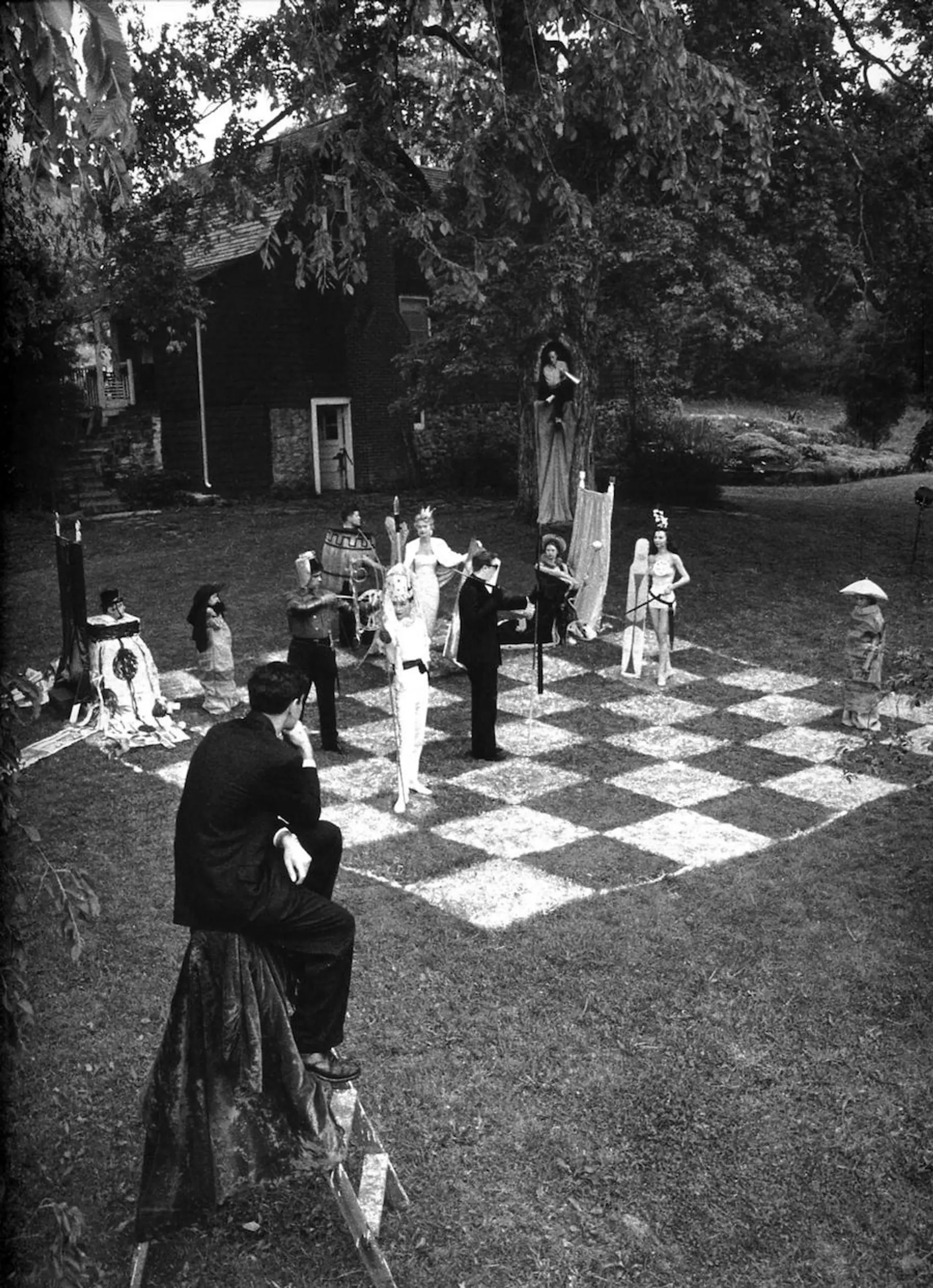 Black-and-white photograph of an outdoor human chess game featuring elaborately dressed participants in a garden setting.