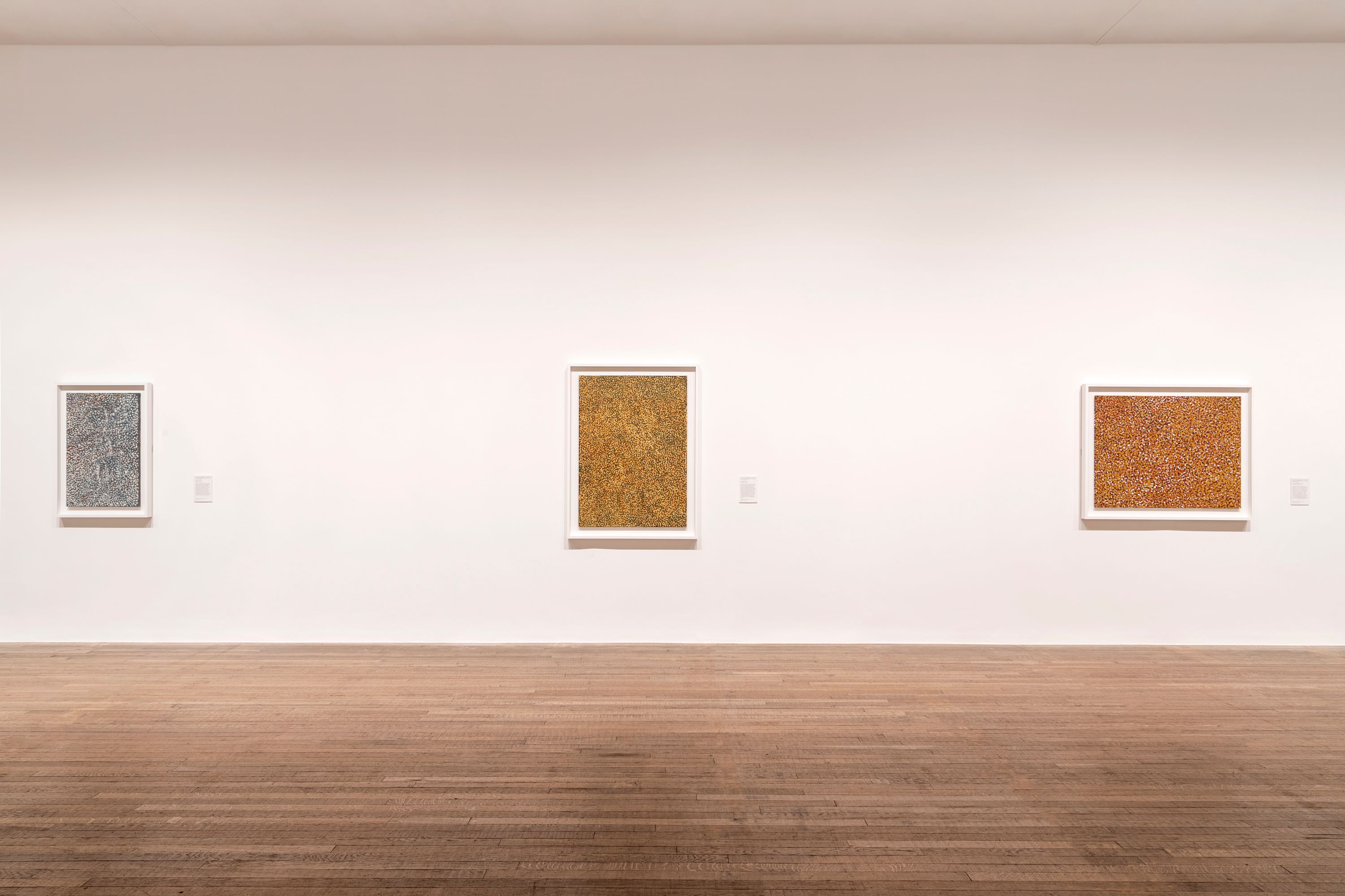 Artworks in various shapes and sizes displayed on a clean, white wall in a gallery with wooden flooring.
