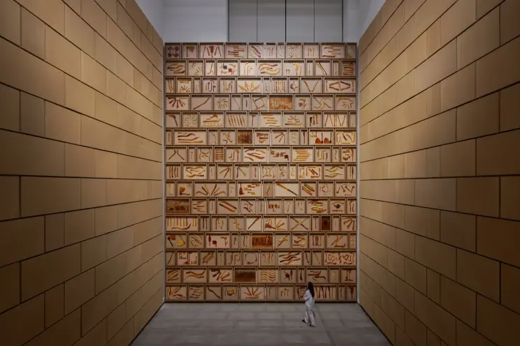 Large wall display of numerous wooden carvings in an enclosed space with a person for scale.