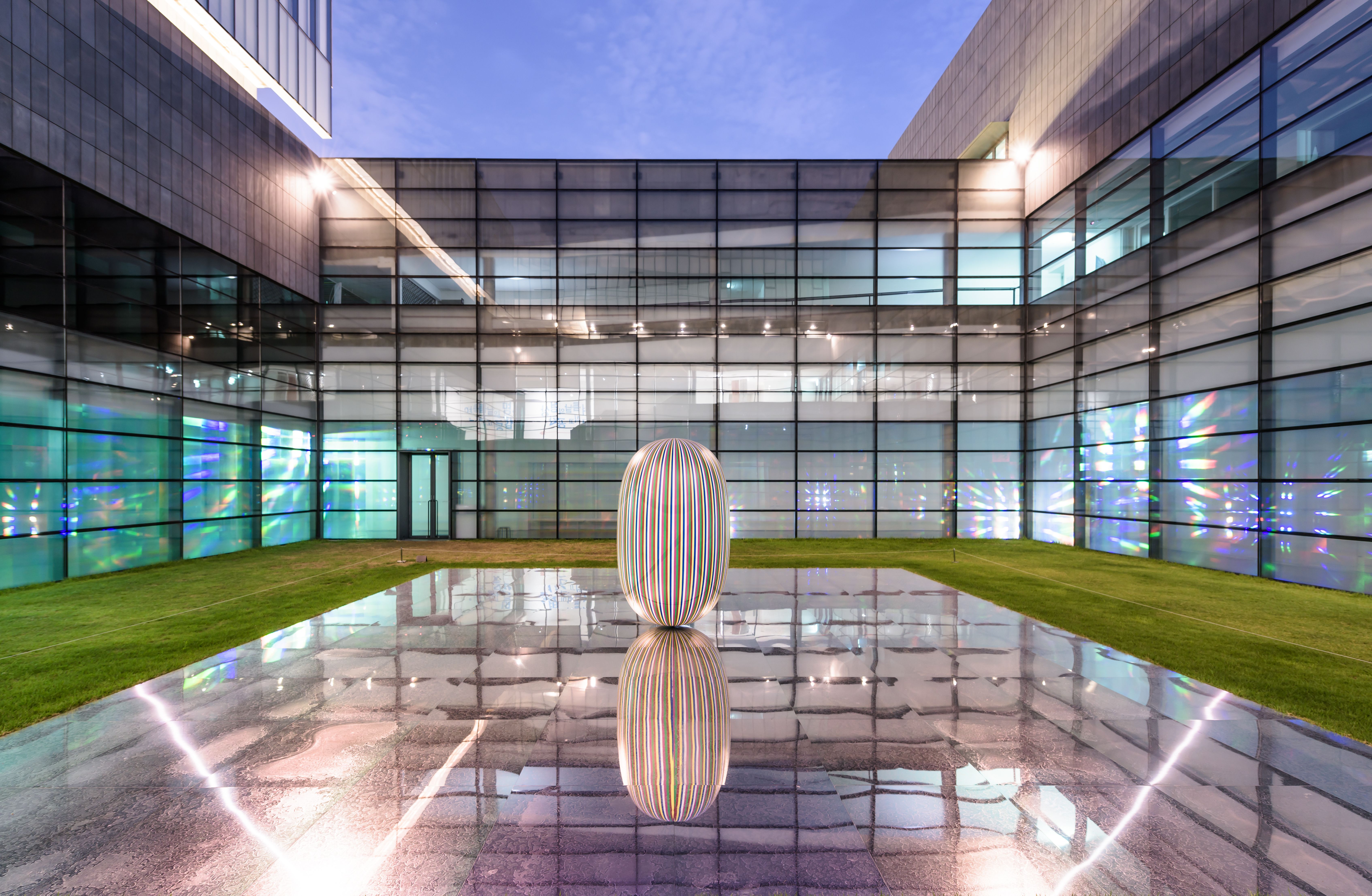 An elliptical sculpture sits on a reflective rectangular surface in the backyard of a building adorned with numerous windows.