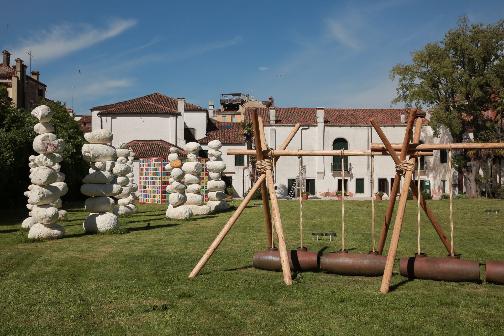 Green lawn before an old building with art installations: stone stacks, colorful squares, and a wooden swing.