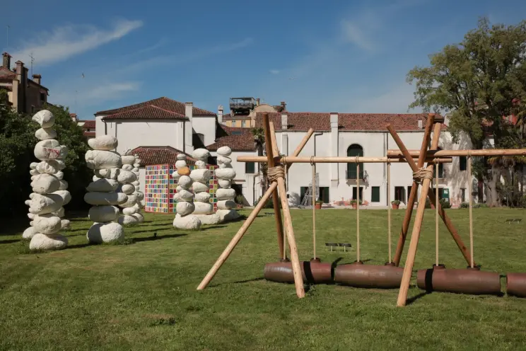 Green lawn before an old building with art installations: stone stacks, colorful squares, and a wooden swing.