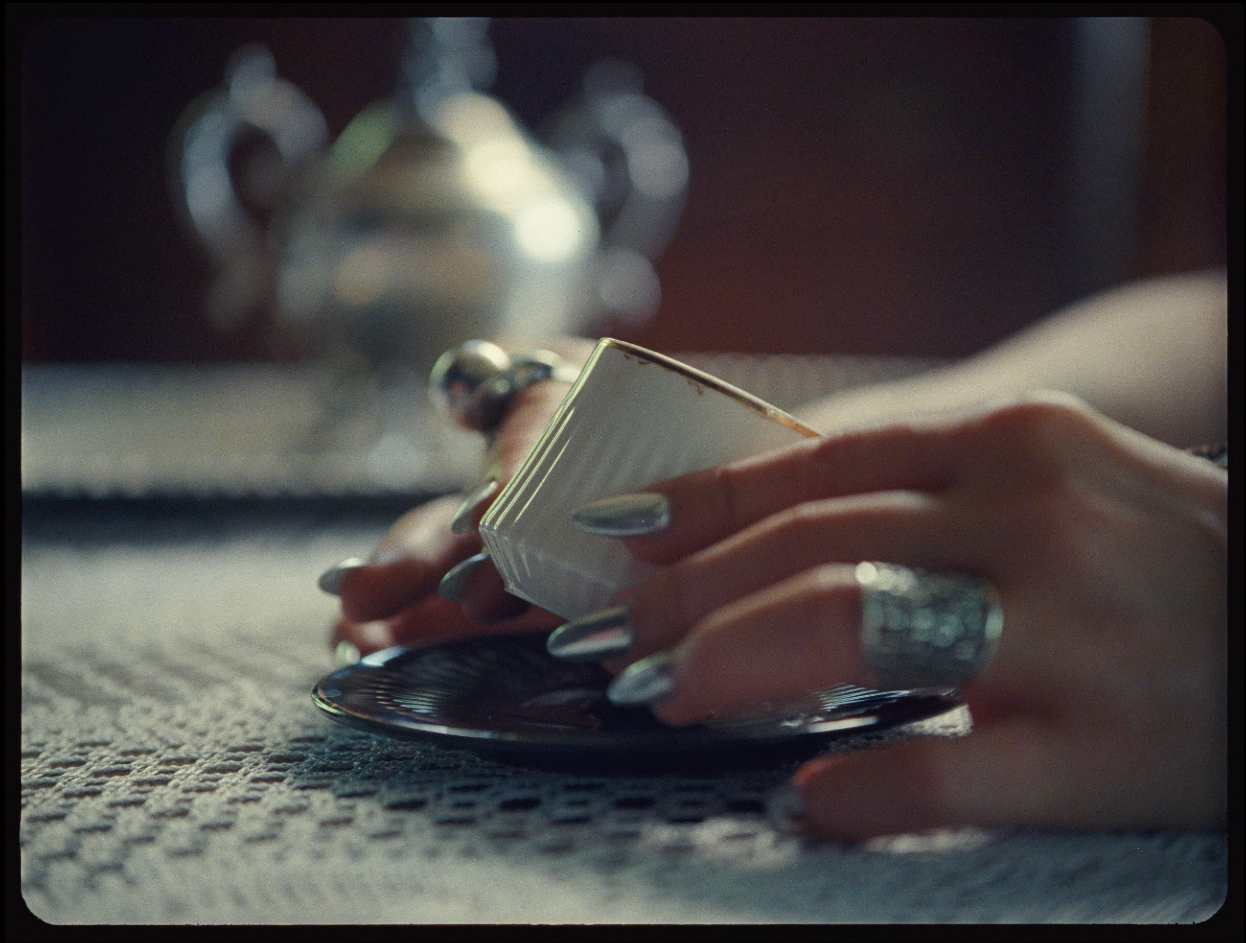 A hand with silver nails holding a small coffee cup over a saucer, set on a lace tablecloth.