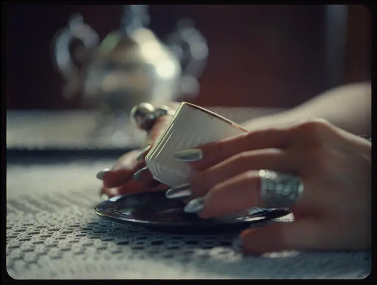 A hand with silver nails holding a small coffee cup over a saucer, set on a lace tablecloth.