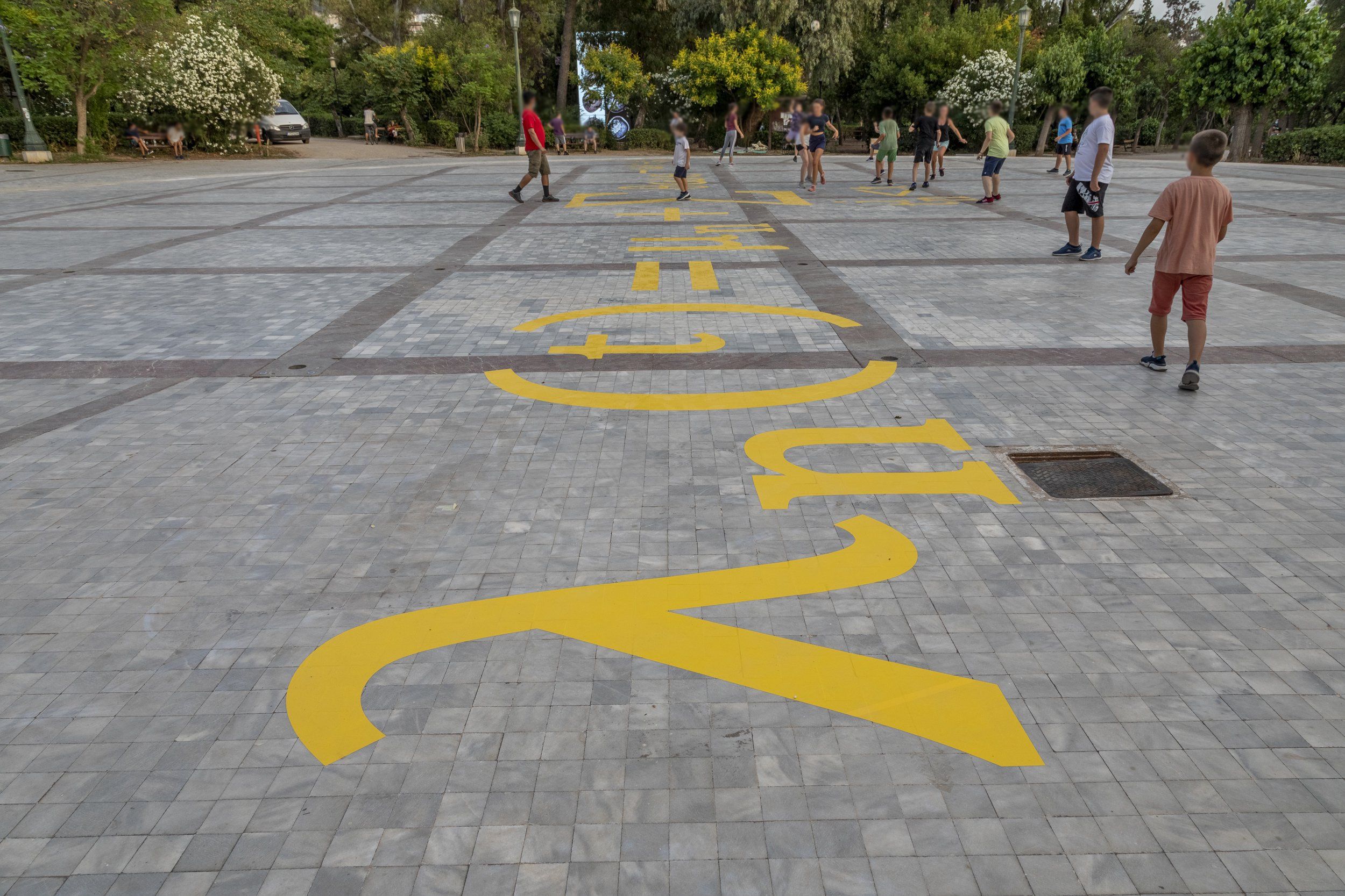 Children playing on a tiled plaza with large yellow mathematical symbols painted on the ground.