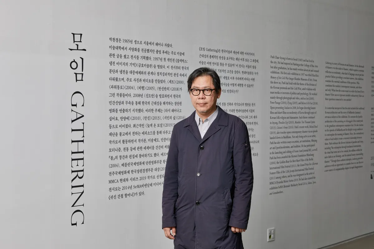 Park Chan-kyong, the artist, stands against a wall with printed text, looking directly at the camera.