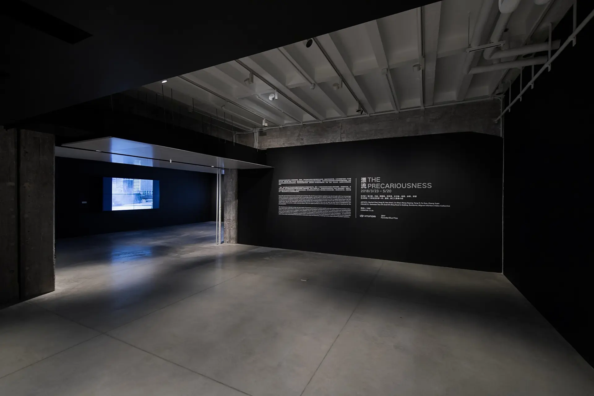 Dark gallery room with a lit text wall and video projection.
