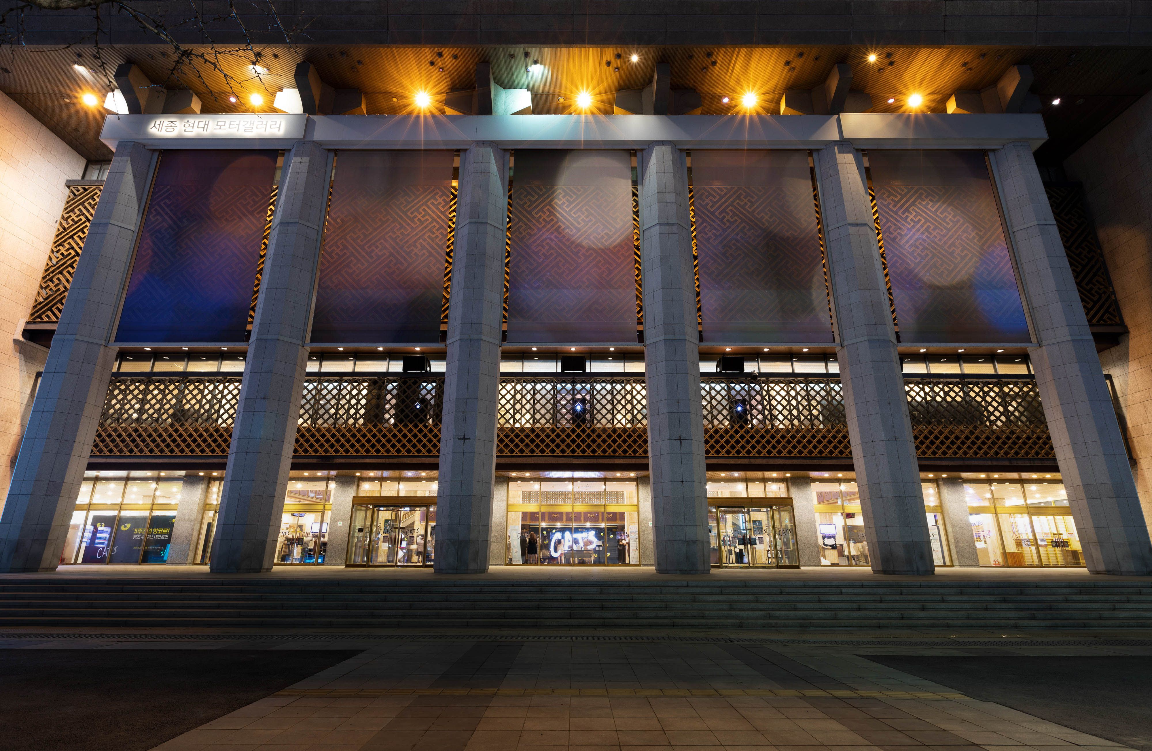Building façade featuring columns, steps and intricate wooden latticework illuminated by yellow lights.